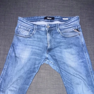 Replay Anbass ljusblå jeans - Säljer ett par Replay Anbass jeans i ljusblå tvätt med klassisk femficksdesign och snygga slitningar. Jeansen har normal passform och raka ben, med dragkedja och knapp i gylfen. Perfekta för dig som gillar stilrena och bekväma jeans.