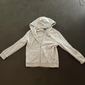 Vit hoodie från Emporio Armani EA7 st 150-155 - Säljer en vit hoodie från Emporio Armani EA7 med dragkedja och huva. Jackan har logga på bröstet och två fickor framtill. Perfekt för dig som gillar stilrena och sportiga plagg. Hoodien är använd två gånger och det finns inga fel med den. Den har storleken 150-155 men passar även 160. Nypris var 700 kr.