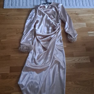 Champagnefärgad långklänning i satin med fjädrar - Säljer en elegant långklänning i champagnefärgad satin med långa ärmar och fjäderdetaljer vid ärmsluten. Klänningen har drapering framtill och knappar längs ryggen. Perfekt för dig som vill sticka ut på festen!