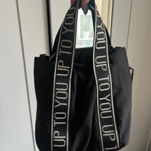Svart axelväska med textade band - Säljer en svart axelväska med breda axelband där det står 'UP TO YOU' i vitt. Väskan har dragkedja upptill och är rymlig med flera fack. Perfekt för dig som vill ha en stilren men ändå trendig accessoar. Kan bäras med både handtag och axelband. Aldrig använt så säljer. Skicka för fler bilder💞