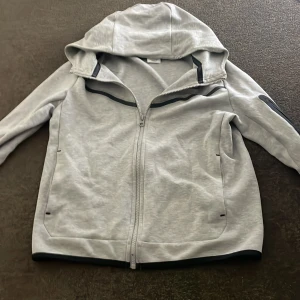 Ljusgrå niketech från Nike st 160 - Snygg ljusgrå niketech från Nike med hel dragkedja och huva. Hoodien har svarta detaljer, fickor framtill och en extra ficka med dragkedja på ärmen. Perfekt för en sportig stil. Cirka st 160
