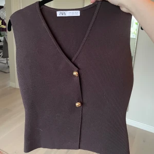 Brun stickad väst från Zara - Säljer en brun stickad väst från Zara med v-ringning och två dekorativa guldknappar framtill. Västen har en enkel, figursydd passform och är ärmlös