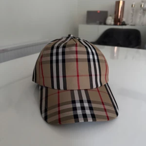 Rutig keps från Burberry - Snygg keps från Burberry med klassiskt rutigt mönster i beige, svart, rött och vitt. Helt ny! Justerbar baktill med plastspänne och tydlig Burberry London England-logga. Perfekt accessoar för en trendig look. Inga returer. 