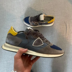 Grå och blå sneakers med gula detaljer från Philippe models  - Snygga sneakers från Philippe Model i grå mocka och blå textil med gula detaljer vid hälen. Skorna har vit sula, snörning och en ikonisk sköld på sidan. Perfekta för en trendig och sportig look. Nypris: 3000kr mitt pris: 1000kr. Storlek 42 passar 43