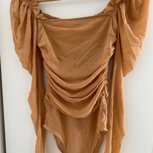 Beige omlott bodysuit med puffärmar - Trendig beige bodysuit med långa puffärmar och snygga draperingar framtill och baktill. Omlott-detalj och figurnära passform ger en cool och modern look. Perfekt för att lyfta vilken outfit som helst.