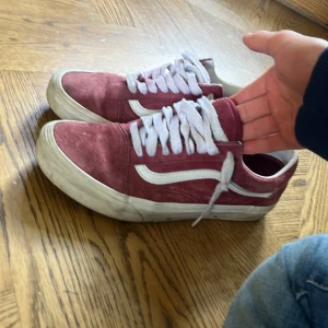 Vinröda Vans Old Skool sneakers - Säljer ett par klassiska Vans Old Skool sneakers i vinrött med vit sula och vit sidoremsa. Skorna har snörning och är tillverkade i mocka och syntetmaterial. Perfekta för en avslappnad och stilren look.