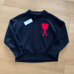 Svart stickad tröja från Ami Paris - Svart stickad tröja från Ami Paris med ett rött hjärta broderat på bröstet. Tröjan har ribbade muddar och rund hals. Perfekt för dig som gillar stilrena detaljer.