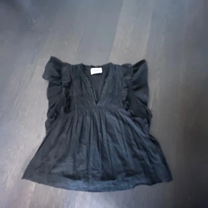 Svart blus med volang från Neo Noir - Säljer en svart blus från Neo Noir med v-ringning och volangdetaljer längs axlar och bröst. Blusen har en luftig passform och är perfekt för dig som gillar en stilren men ändå söt look.