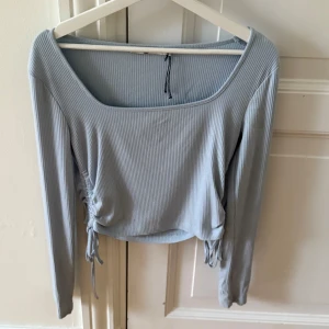 Ljusblå ribbad croppad topp från NA-KD - Säljer en ljusblå croppad topp från NA-KD med långa ärmar och ribbad struktur. Toppen har snygga knyt- och rynkdetaljer i sidorna och en bred, fyrkantig ringning. Perfekt för att lyfta din vardagsstil!