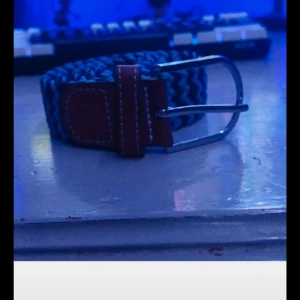 Blått flätat bälte med skinn-detaljer - Säljer ett snyggt blått bälte med flätad design och bruna skinn-detaljer vid spännet. Bältet har ett klassiskt silverfärgat spänne och passar perfekt till jeans eller chinos.