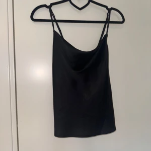 Svart satinlinne med smala axelband - Säljer ett svart linne i satin med smala, justerbara axelband som korsas i ryggen. Linnet har en enkel och stilren design med lätt glansigt material. Perfekt att styla till olika outfits.