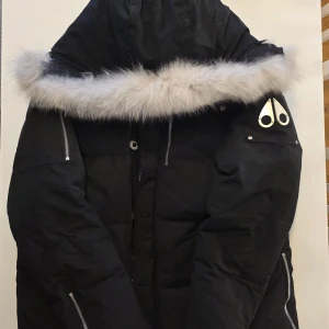 Svart dunjacka med päls från Moose Knuckles - Säljer en svart dunjacka från Moose Knuckles med avtagbar huva och lyxig pälskant i vitt. Jackan har flera dragkedjor, fickor, metallogo på armen och en klassisk pufferlook. Perfekt för kalla dagar och riktigt snygg. Enligt alla reviews och recensioner är det den varmaste jackan någonsin – perfekt för iskalla vinterdagar. Det står 2XL i jackan, men den passar som en vanlig storlek L med tanke på att man bär en tröja under  **säljs för billigt eftersom det inte är säsong