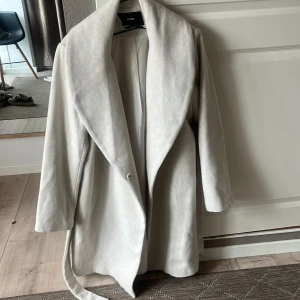Ljusbeige kappa från BikBok - Säljer en stilren ljusbeige kappa från BikBok i storlek XS. Kappan har bred krage, knytbälte i midjan och långa ärmar. Perfekt för dig som vill ha en klassisk och elegant look.