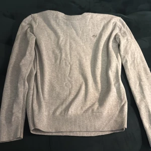 Ljusgrå tröja från GANT - Säljer en stilren ljusgrå tröja från GANT med broderad logga på bröstet. Tröjan har rund halsringning, långa ärmar och ribbade muddar vid ärmslut och nederkant. Perfekt för en clean och klassisk look.