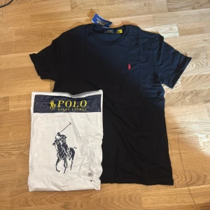 Svart t-shirt från Polo Ralph Lauren - Svart t-shirt från Polo Ralph Lauren med klassisk passform och rund hals. T-shirten har en liten röd broderad logga på bröstet och är tillverkad i mjuk bomull. Perfekt för en stilren och enkel look.