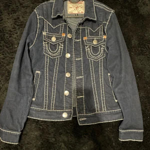 True religion jacket - Säljer en mörkblå jeansjacka från True Religion med kontrasterande vita sömmar och silverfärgade knappar. Jackan har två bröstfickor med lock och knapp samt klassisk krage. Perfekt för dig som vill ha en snygg och tidlös denimlook.