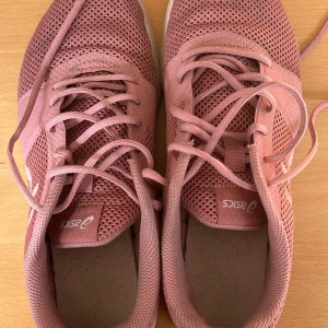 Rosa sneakers från Asics - Säljer ett par rosa Asics sneakers med mesh-överdel och vita detaljer. Skorna har snörning och platt sula, perfekta för träning eller vardagsbruk. Tillverkade i syntetmaterial med EVA-sula för extra komfort.