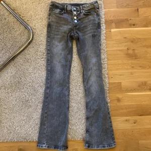 Grå bootcut jeans med knappar - Säljer ett par grå bootcut jeans med hög midja och knappgylf. Jeansen har klassiska fem fickor och bälteshällor. Snygg tvätt och passar perfekt till en trendig vardagslook.