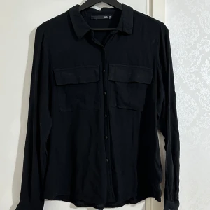 Svart skjorta från 157 - Säljer en svart långärmad skjorta från 157 i storlek XS. Skjortan har klassisk krage, två bröstfickor med lock och knäppning framtill. Perfekt för en stilren och enkel look.
