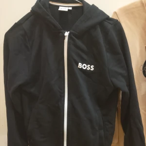 Svart hoodiejacka från Hugo Boss - Svart hoodiejacka från Boss med vit dragkedja och logga på bröstet. Jackan har huva och är långärmad, perfekt för en avslappnad stil.