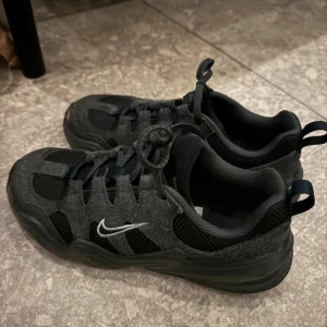 Svarta Nike sneakers med mesh och mockadetaljer - Säljer ett par svarta Nike sneakers med meshpaneler och mockadetaljer. Skorna har snörning, rund tå och en platt sula. Swoosh-loggan syns på sidan i vitt. Perfekta för vardagsbruk och streetstyle.