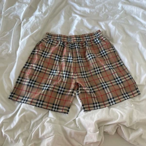 Rutigta badshorts från Burberry - Snygga badshorts från Burberry i klassiskt beige rutigt mönster med inslag av svart, vitt och rött. Shortsen har elastisk midja och innerbyxa i mesh för extra komfort. Perfekta för sommardagar vid stranden eller poolen.