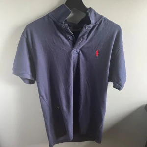 Mörkblå pikétröja från Ralph Lauren - Säljer en klassisk mörkblå pikétröja från Ralph Lauren med korta ärmar och röd broderad logga på bröstet. Tröjan har normal passform och är tillverkad i mjuk bomull. Perfekt för en stilren och avslappnad look.