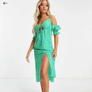 Grön blommig klänning med volang och slits - Snygg grön klänning med vitt blommönster, volangdetaljer på ärmarna och knytning framtill. Klänningen har axelband, korta puffärmar och en hög slits framtill. Perfekt för sommarens alla tillfällen.