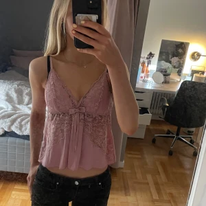 Rosa spetsig linnetopp med rosett - Söt ljusrosa linnetopp med spetsdetaljer och rosett framtill. Toppen har v-ringning och tunna axelband. Perfekt för varma dagar eller som ett lager under en kofta.