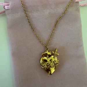 Taira Hjärthalsband  - Unikt halsband i guld med ett detaljerat hjärthänge dekorerat med rosor och kors. Kedjan är tunn och elegant, perfekt för att ge en personlig touch till din stil. Nytt skick och köpte precis men det var inte riktigt min stil❤️