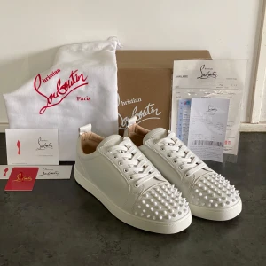 Christian Louboutin Vita läder - Säljer ett par vita sneakers från Christian Louboutin med ikoniska röda sulor och silverfärgade nitar på tån. Skorna har snörning och är tillverkade i skinn. Perfekta för dig som vill sticka ut med en lyxig och edgy stil.