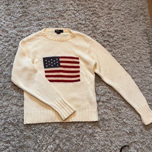 Vit stickad tröja med amerikansk flagga från Polo Ralph Lauren - Superfin stickad tröja från Ralph Lauren med broderad USA-flagga på framsidan. Äldre vintage-modell, tillverkad i Hongkong – troligen från 90-talet. Tjock, mjuk bomull i off-white med mörkblått och vinrött motiv.  Skick: Mycket bra! En liten fläck på ryggen som knappt syns (bild finns).  Perfekt för dig som gillar vintage eller preppy stil!