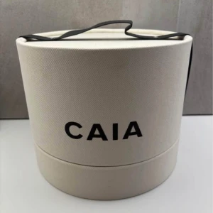 Förvaringsbox från CAIA - Säljer en rund, vit sminkborstbox från CAIA med svart text och lock. Perfekt för att förvara sminkborstar eller andra sminkprodukter. Snygg och stilren design som passar på sminkbordet.