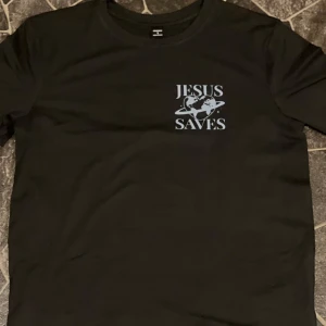 Svart t-shirt med tryck 'Jesus Saves' - Svart t-shirt med vitt tryck på bröstet och ryggen där det står 'Jesus Saves' tillsammans med en jordglob i hjärtform och bibelcitat. Klassisk rund hals och korta ärmar. Perfekt för dig som vill bära ett tydligt statement.
