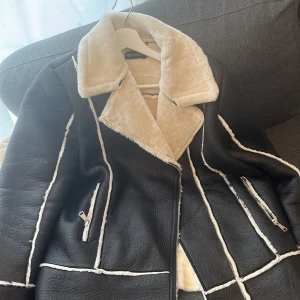 Svart pälsjacka med vit fuskpäls från PrettyLittleThing - Säljer en svart aviator jacka från PrettyLittleThing med vit fuskpäls på krage och insida. Jackan har dragkedja framtill och två fickor med dragkedja. Snygga kontrastsömmar i vitt ger en trendig look. Perfekt för höst/vinter aldrig använd. 