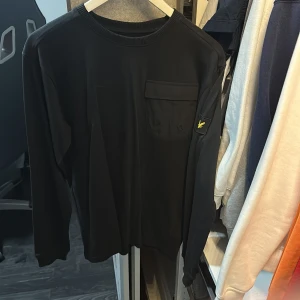 Svart långärmad tröja från Lyle & Scott Junior - Säljer en svart långärmad tröja från Lyle & Scott Junior med rund halsringning och bröstficka. Tröjan har en gul logga på ärmen och är tillverkad i mjuk bomull. Perfekt för en stilren och enkel look.