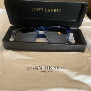 Blå solglasögon från John Henric - Snygga blå solglasögon från John Henric med rundad form och mörka glas. Bågarna är i transparent blå plast och ger en modern och stilren look. Perfekta för soliga dagar och levereras med originalfodral och putsduk.