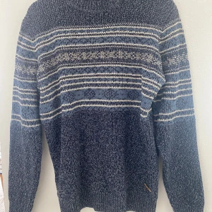 Blå stickad tröja med mönster från Indicode - Säljer en blå stickad tröja från Indicode i storlek Large. Tröjan har ett randigt mönster i blått, vitt och beige, samt rund halsringning och långa ärmar. Perfekt för kyligare dagar och enkel att matcha med jeans.