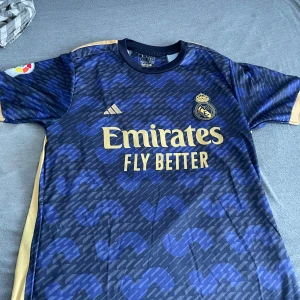 Real Madrid fotbollströja Vini Jr 20 - Säljer en blå och guld Real Madrid fotbollströja från Adidas med Vini Jr och nummer 20 på ryggen. Tröjan har korta ärmar, gulddetaljer och klubbmärke samt sponsortryck på bröstet. Perfekt för fans av Real Madrid och Vini Jr.