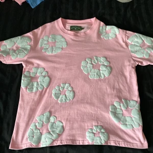 Rosa t-shirt med mintgrönt mönster Denim Tears - Unik rosa t-shirt från Denim Tears med mintgröna, vadderade cirkelmönster över hela plagget. Oversized fit och tillverkad i mjuk bomull. Perfekt statement-piece för dig som vill sticka ut med cool design och färg.