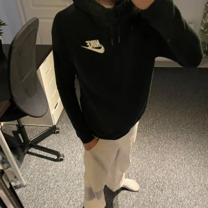 Svart Nike hoodie XS - Svart hoodie från Nike i storlek XS med klassisk vit logga på bröstet. Tröjan har huva med snörning och är långärmad. Perfekt för chill dagar eller när du vill ha en enkel och stilren look.