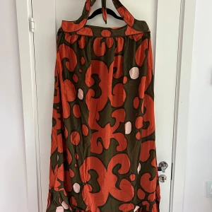 Marimekko x H&M långklänning 42 - Snygg långklänning från Marimekko x H&M med stort grafiskt mönster i orange, olivgrönt och ljusrosa. Klänningen har halterneck med knyt i nacken och dragkedja bak. Luftig modell i bomull, perfekt för dig som gillar statement-plagg.
