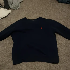 Helt ny Mörkblå tröja från Polo Ralph Lauren andvänd en gång - Mörkblå crewneck-tröja från Polo Ralph Lauren med klassisk röd broderad logga på bröstet. Tröjan har rund halsringning och långa ärmar, perfekt för en clean och stilren look. Tillverkad i mjuk bomull för skön komfort.