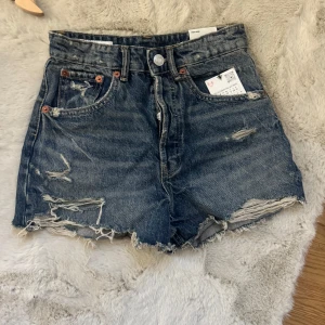 Högmidjade jeansshorts från Zara - Snygga högmidjade jeansshorts från Zara i mörkblå denim med slitna detaljer och fransig kant. Klassisk femficksmodell med knapp och dragkedja. Perfekta för en chill sommarlook och passar till allt från sneakers till boots.
