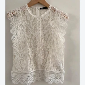 Vit spetsblus från Zara - Superfin vit blus från Zara i spets med blommigt mönster och snygga volangdetaljer längs ärmhålen. Blusen är ärmlös och har rund halsringning samt dragkedja bak. Perfekt för en fräsch och romantisk look.