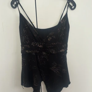 Svart glittrig topp med  blommor - Supersöt svart topp med tunna axelband och omlottliv. Toppen har glittriga blommönster i silver och en flowy kjol. Perfekt för dig som vill sticka ut med lite extra glitter. Materialet känns lätt och luftigt.