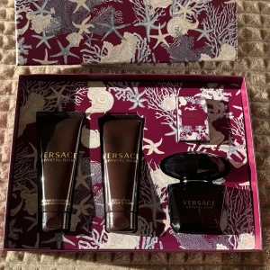 Versace Crystal Noir presentbox - Lyxig presentbox från Versace Crystal Noir med parfym (90 ml), miniparfym (5 ml), duschgel (100 ml) och body lotion (100 ml). Allt kommer i en snygg mörklila ask med havsinspirerat mönster och gulddetaljer. Flaskorna är mörka med guldtext och elegant design.