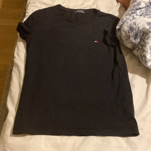 Svart t-shirt från Tommy Hilfiger - Svart t-shirt från Tommy Hilfiger med klassisk rund hals och liten broderad logga på bröstet. Enkel och stilren design, perfekt till jeans eller shorts. Tillverkad i mjuk bomull för skön känsla hela dagen.