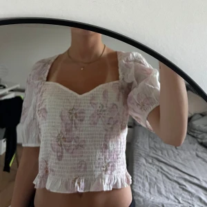 Vit blommig blus med puffärm - Supersöt blus i vitt med ljusrosa blommor. Toppen har puffiga korta ärmar, söt volangkant nertill och en hjärtformad urringning. Älskar denna top men den är tyvär för stor och där av sitter inte so bra, men skriv om ni vill ha fler bilder.