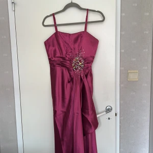Lång rosa balklänning/fest klänning. Stl: Small  - Snygg långklänning i ceriserosa (mörkrosa) med smala axelband och glansigt satintyg. Framtill finns en dekoration med pärlor och stenar som ger extra bling. Klänningen är helt ny, oanvänd!  Perfekt för bal eller festkväll!